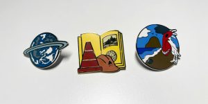 Photo of Mysterium 2025 pin set with a planet for Todelmer on the left, a Relto book with DRC helmet and cone in the middle, and piranha chicken on a beach on the right.